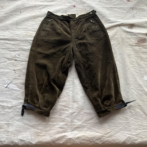 K&ouml;nnte beinhalten: Dunkel olivgr&uuml;ne Cordhose mit hohem Bund und verk&uuml;rzter L&auml;nge. Merkmale sind Vordertaschen, Knopf- und Rei&szlig;verschluss sowie verstellbare Kn&ouml;chelriemen. Die Hose ist aus einem weichen, strukturierten Stoff gefertigt.