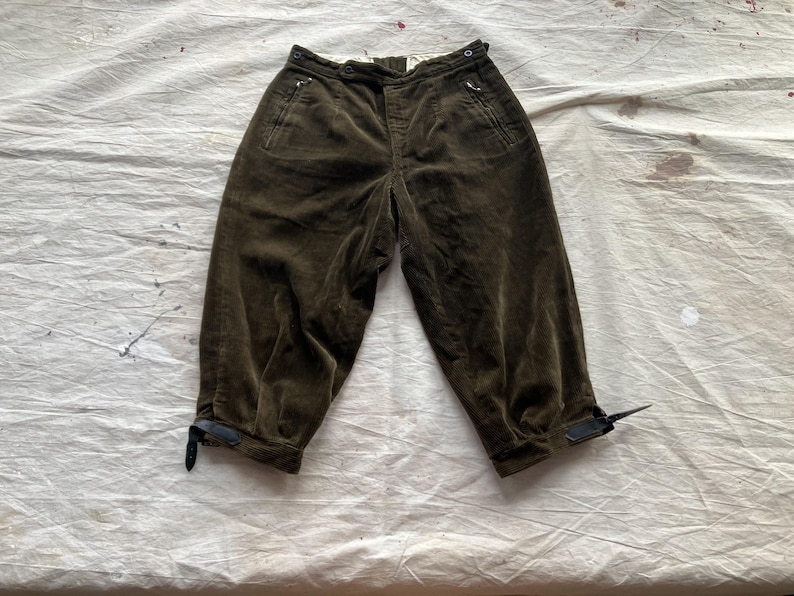 K&ouml;nnte beinhalten: Dunkel olivgr&uuml;ne Cordhose mit hohem Bund und verk&uuml;rzter L&auml;nge. Merkmale sind Vordertaschen, Knopf- und Rei&szlig;verschluss sowie verstellbare Kn&ouml;chelriemen. Die Hose ist aus einem weichen, strukturierten Stoff gefertigt.