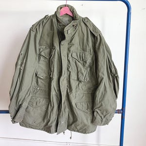 Könnte beinhalten: Olivgrüne Jacke im Militärstil mit mehreren Fronttaschen und hohem Kragen. Die Jacke hängt an einem rosa Kleiderbügel, der an einem blauen Metallgestell befestigt ist. Die Jacke hat eine Knopfleiste und Schulterklappen.