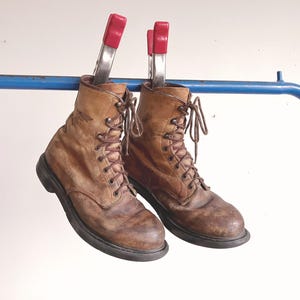 Könnte beinhalten: Ein Paar abgenutzte, hellbraune Schnürstiefel aus Leder mit dunkelbraunen Schnürsenkeln und schwarzen Sohlen. Die Stiefel hängen an einer blauen Metallstange und sind mit zwei silbernen Metallklemmen mit roten Kunststoffgriffen befestigt.