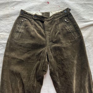 K&ouml;nnte beinhalten: Nahaufnahme einer olivgr&uuml;nen Cordhose. Die Hose hat eine hohe Taille, einen Knopfverschluss und Seitentaschen. Der Stoff hat eine gerippte Textur. Die Hose liegt flach auf einer wei&szlig;en Oberfl&auml;che.