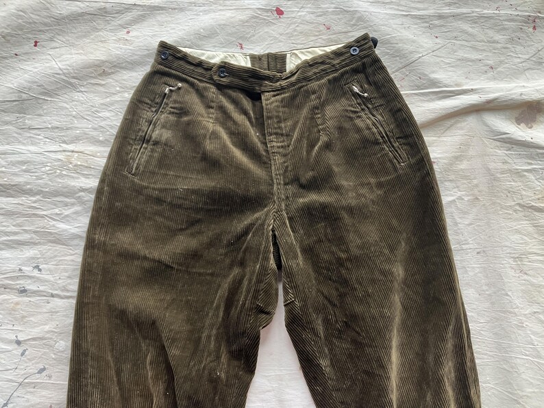 K&ouml;nnte beinhalten: Nahaufnahme einer olivgr&uuml;nen Cordhose. Die Hose hat eine hohe Taille, einen Knopfverschluss und Seitentaschen. Der Stoff hat eine gerippte Textur. Die Hose liegt flach auf einer wei&szlig;en Oberfl&auml;che.