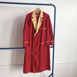 May include: A red robe with a yellow collar and trim hangs on a wooden hanger. The robe has a double-breasted design with buttons and pockets. The robe is made of a lightweight fabric and appears to be vintage.