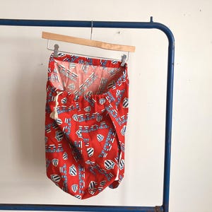 May include: Red laundry bag with a repeating pattern of white stars and blue shield-like shapes. The bag hangs from a wooden hanger on a blue metal rack. A white drawstring is visible.