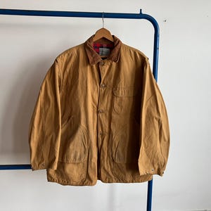 Può includere: Giacca color marrone con colletto in velluto a coste e fodera a quadri rossi e neri. La giacca ha quattro tasche anteriori ed è abbottonata. È appesa a una gruccia di legno.