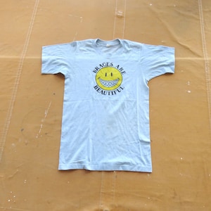 Puede incluir: Camiseta azul claro con una cara sonriente amarilla que lleva brackets y el texto "Braces Are Beautiful".
