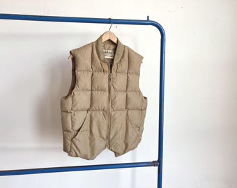 Könnte beinhalten: Eine beige Steppweste mit quadratischem Steppmuster, durchgehendem Frontreißverschluss und geripptem Kragen. Das Kleidungsstück verfügt über zwei Vordertaschen und ein Cabela's Markenetikett im inneren Nackenbereich.