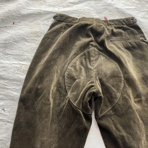 K&ouml;nnte beinhalten: Nahaufnahme einer olivgr&uuml;nen Cordhose. Die Hose hat ein einzigartiges Design mit einem kreisf&ouml;rmigen Aufn&auml;her am Ges&auml;&szlig;. Der Stoff hat vertikale Rippen, und es gibt einen Knopf und eine Tasche.