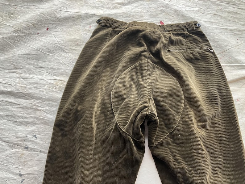 K&ouml;nnte beinhalten: Nahaufnahme einer olivgr&uuml;nen Cordhose. Die Hose hat ein einzigartiges Design mit einem kreisf&ouml;rmigen Aufn&auml;her am Ges&auml;&szlig;. Der Stoff hat vertikale Rippen, und es gibt einen Knopf und eine Tasche.
