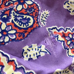 May include: Close-up of a section of fabric with a paisley pattern. The fabric is a light purple colour with a repeating design in red, blue, and yellow. There is a tear in the fabric.