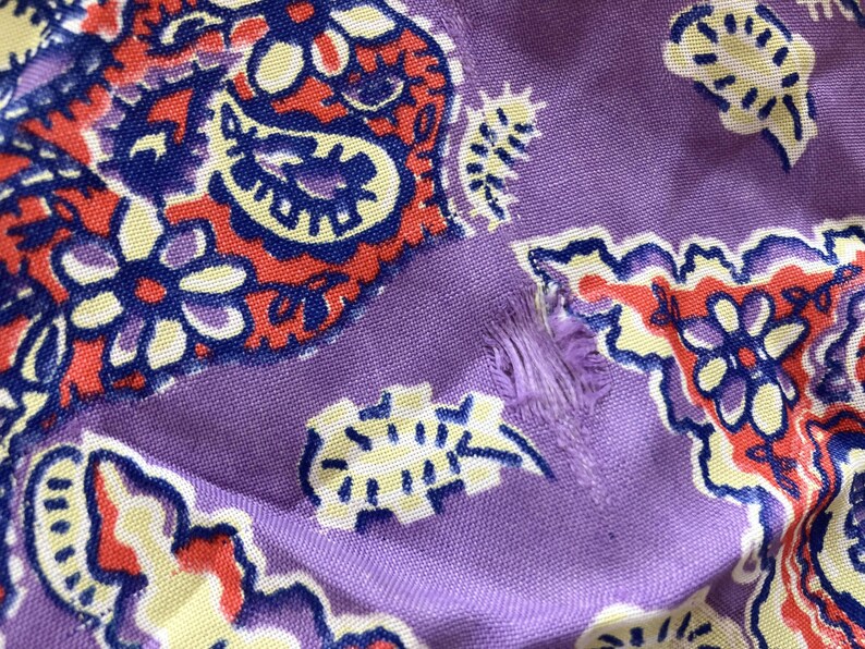 May include: Close-up of a section of fabric with a paisley pattern. The fabric is a light purple colour with a repeating design in red, blue, and yellow. There is a tear in the fabric.