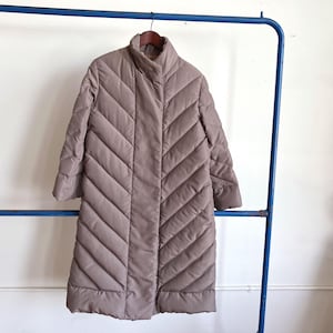 May include: A long, taupe quilted coat with a high collar and button closure. The coat is hanging on a blue metal clothing rack. The coat has a chevron pattern.