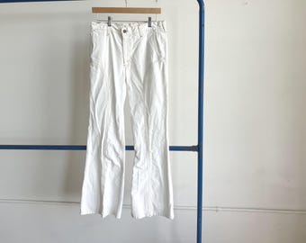 May include: A pair of off-white pants featuring a high-waisted design, a button and zipper fly closure, and prominent vertical seam detailing running down the front of each leg. The pants have a flared silhouette and are hanging on a wooden hanger.