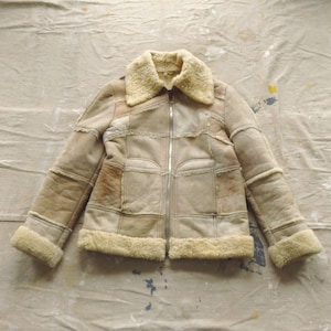 May include: A beige suede jacket with a faux fur collar and zipper closure. The jacket has a patchwork design and is made of multiple pieces of suede sewn together.