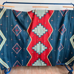 May include: A woven textile with a geometric pattern. The rug features a central red diamond shape flanked by blue sections with diamond and cross motifs. The edges have a yellow and green zigzag design, with fringe.