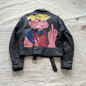 Op de afbeelding: Zwarte leren jas met een kleurrijke cartoonafbeelding op de rug. De afbeelding toont een roze personage dat de middelvinger opsteekt. De jas heeft een gespriem en een cropped ontwerp.