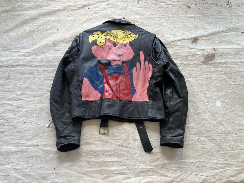Op de afbeelding: Zwarte leren jas met een kleurrijke cartoonafbeelding op de rug. De afbeelding toont een roze personage dat de middelvinger opsteekt. De jas heeft een gespriem en een cropped ontwerp.