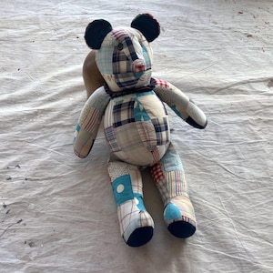 May include: A patchwork teddy bear made from various fabrics in blue, red, and white patterns. The bear features dark blue ears and feet, and a brown body. The bear is positioned on a white surface.