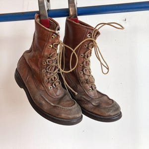 Puede incluir: Un par de botas de cuero marrón con cordones, diseño de puntera tipo mocasín, ojales metálicos y ganchos para los cordones. Las botas tienen un acabado desgastado, suelas de goma marrón oscuro y cuelgan de una barra azul con abrazaderas.