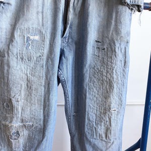 May include: Light blue denim pants with visible wear and tear, featuring patches and distressed areas. The pants have a zipper fly and are displayed on a blue metal rack. The overall look suggests a vintage or workwear style.