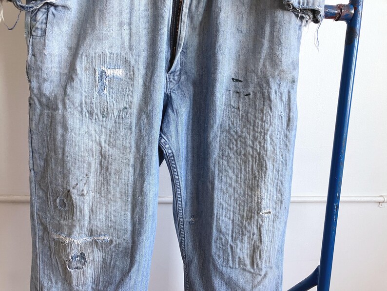 May include: Light blue denim pants with visible wear and tear, featuring patches and distressed areas. The pants have a zipper fly and are displayed on a blue metal rack. The overall look suggests a vintage or workwear style.