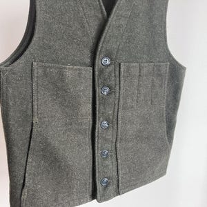 May include: A dark olive green wool vest featuring a V-neckline, a front button closure with five dark buttons, and two large patch pockets on the front. The garment has a structured design with visible seams and a thick, textured fabric construction.