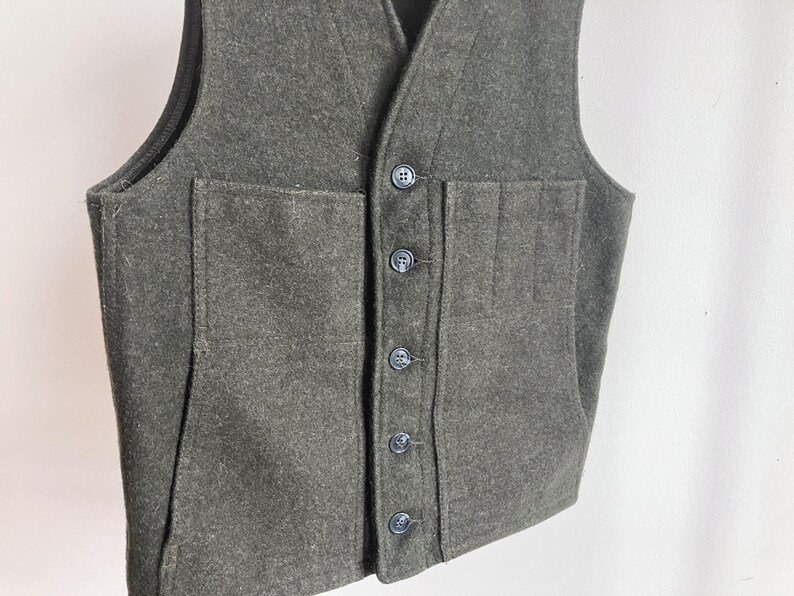 May include: A dark olive green wool vest featuring a V-neckline, a front button closure with five dark buttons, and two large patch pockets on the front. The garment has a structured design with visible seams and a thick, textured fabric construction.