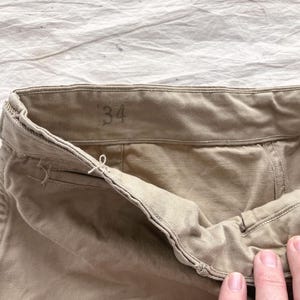 May include: Close-up of khaki-colored pants. The waistband is visible, with the size "34" printed on it. The fabric appears to be a sturdy cotton or blend. The pants are slightly wrinkled, suggesting they have been worn.