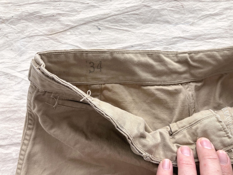 May include: Close-up of khaki-colored pants. The waistband is visible, with the size "34" printed on it. The fabric appears to be a sturdy cotton or blend. The pants are slightly wrinkled, suggesting they have been worn.