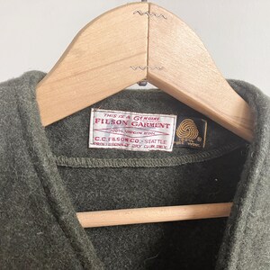 May include: An olive green wool vest featuring a white rectangular label with red text reading This is a Genuine Filson Garment, 100% Virgin Wool, C.C. Filson Co. Seattle, Professionally Dry Clean Only, and a small black tag with a gold wool symbol.