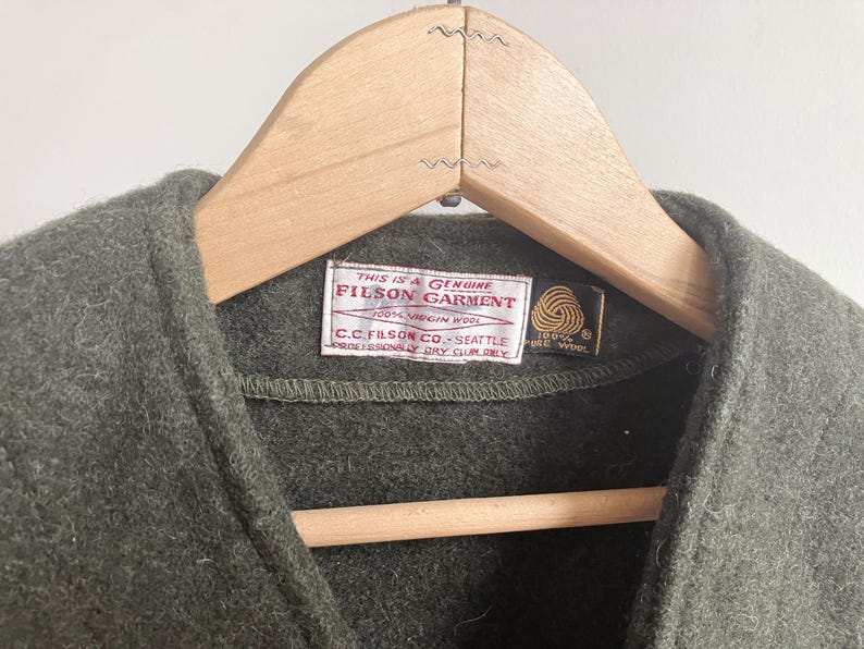 May include: An olive green wool vest featuring a white rectangular label with red text reading This is a Genuine Filson Garment, 100% Virgin Wool, C.C. Filson Co. Seattle, Professionally Dry Clean Only, and a small black tag with a gold wool symbol.