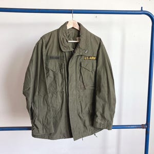 May include: Olive green military jacket with multiple pockets and a collar. The jacket has a "U.S. ARMY" patch and a name patch that reads "NELSON". The jacket is hanging on a wooden hanger.