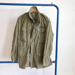 May include: An olive green military-style jacket with multiple front pockets and a button-up closure. The jacket is hanging on a blue metal clothing rack. The jacket has a collared neckline and long sleeves.