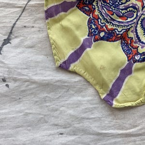May include: A fragment of patterned fabric with a yellow base and purple stripes. The fabric displays a detailed paisley design in purple, red, and blue. The fabric rests on a white surface with grey paint splatters.