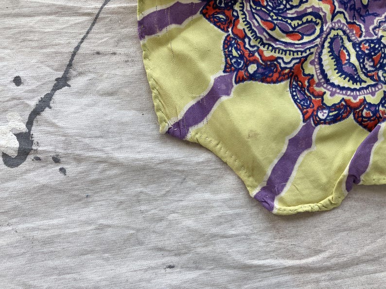 May include: A fragment of patterned fabric with a yellow base and purple stripes. The fabric displays a detailed paisley design in purple, red, and blue. The fabric rests on a white surface with grey paint splatters.