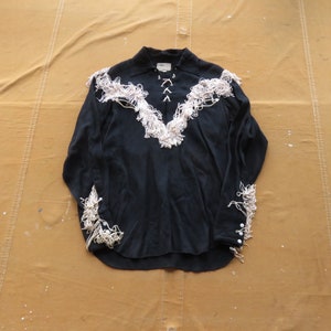 May include: A black long-sleeved shirt with white fringe trim around the collar, cuffs, and front. The shirt has a button-up closure and a pointed collar.