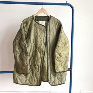 May include: An olive green quilted liner jacket featuring a wave-patterned stitch design, long sleeves, and a collarless neckline. The jacket has a front opening with visible interior tag details and is constructed from a lightweight nylon material.