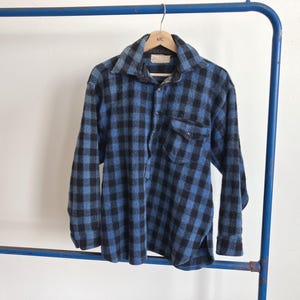 May include: A blue and black plaid flannel shirt with long sleeves hangs on a wooden hanger. The shirt has a button-down front, a collar, and a chest pocket. The shirt is displayed on a blue metal rack against a white background.