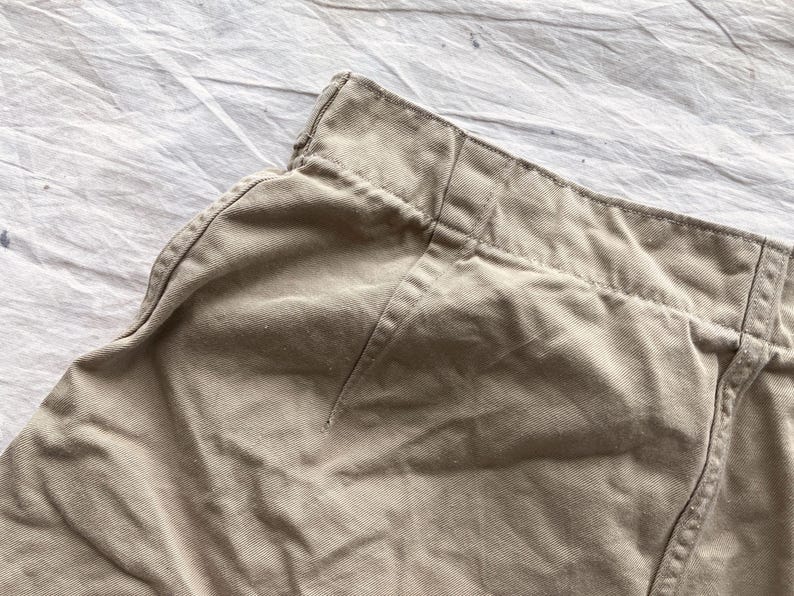 May include: Close-up of tan-colored fabric, possibly part of a garment. The fabric appears to be a sturdy material, with visible stitching along the seams. The image focuses on the waistband and a portion of the garment's side, showcasing the texture and construction.