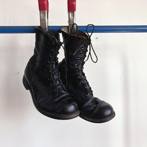 May include: A pair of black leather lace-up combat boots with rounded toes and black laces. The boots are suspended from a blue horizontal bar by two metal clamps with red handles, showing the exterior construction and lacing system.