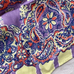 May include: Close-up of a fabric with a paisley pattern in shades of purple, yellow, orange, and blue. The design features floral and teardrop shapes. The fabric has a soft, slightly wrinkled texture.