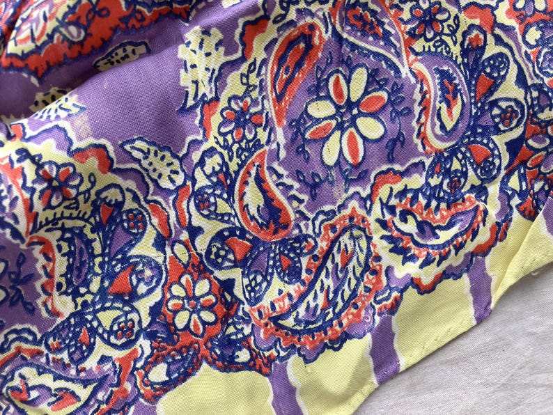 May include: Close-up of a fabric with a paisley pattern in shades of purple, yellow, orange, and blue. The design features floral and teardrop shapes. The fabric has a soft, slightly wrinkled texture.