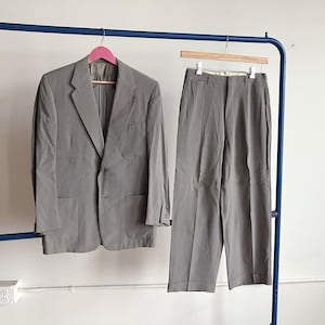 May include: A light gray suit, including a blazer and matching trousers, hangs on a blue clothing rack. The blazer has a two-button closure and two front pockets. The trousers are pleated and have a straight leg.
