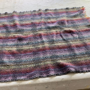 May include: A hand-knitted blanket with a colorful striped pattern in shades of purple, pink, blue, and gold. The blanket has a textured design and fringed edges. This cozy blanket is perfect for adding warmth and style to any room.