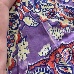 May include: Close-up of a section of fabric with a paisley pattern. The design features a mix of colours, including purple, red, yellow, and blue. The fabric appears to be soft and lightweight, with a slight sheen. The pattern is intricate and detailed.