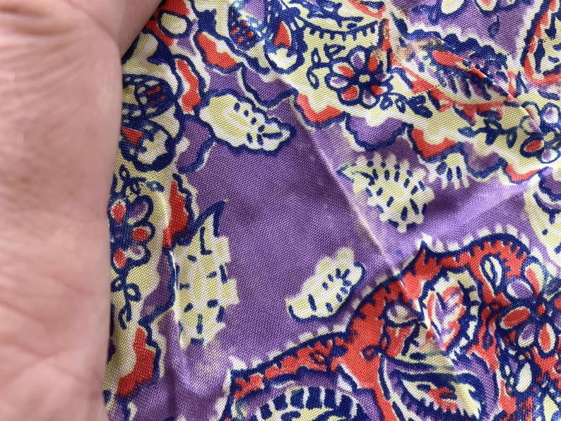 May include: Close-up of a section of fabric with a paisley pattern. The design features a mix of colours, including purple, red, yellow, and blue. The fabric appears to be soft and lightweight, with a slight sheen. The pattern is intricate and detailed.