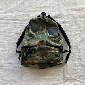 May include: Camouflage-patterned backpack with black shoulder straps and accents. The backpack features a front pocket and two black patches on the front. The bag is made of a durable material and is designed for carrying items.