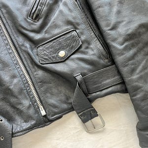 Op de afbeelding: Close-up van een zwarte leren jas met een zilveren rits, een klein zakje met een knoop en een riem met een zilveren gesp. De jas heeft een klassieke biker-stijl.