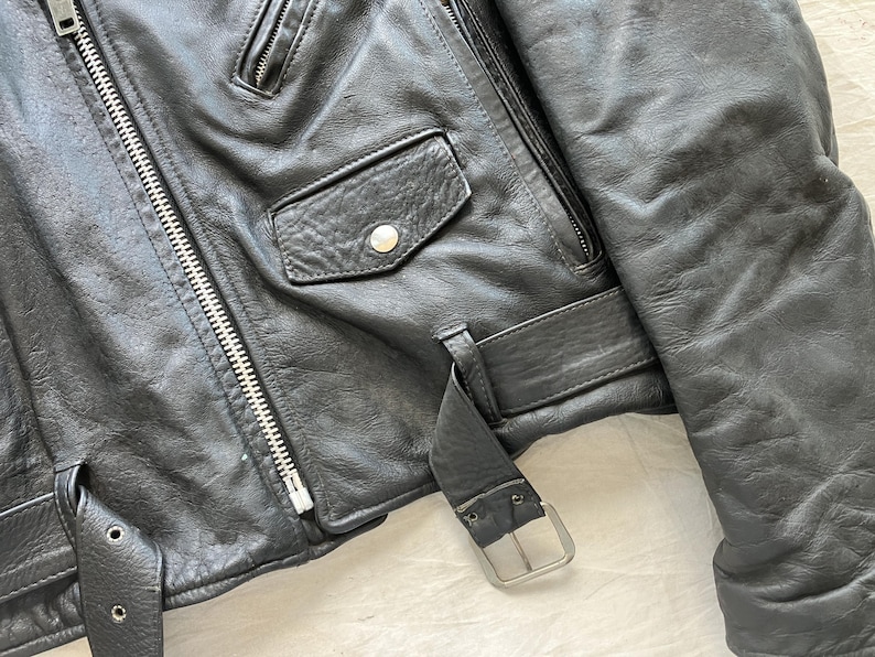 Op de afbeelding: Close-up van een zwarte leren jas met een zilveren rits, een klein zakje met een knoop en een riem met een zilveren gesp. De jas heeft een klassieke biker-stijl.