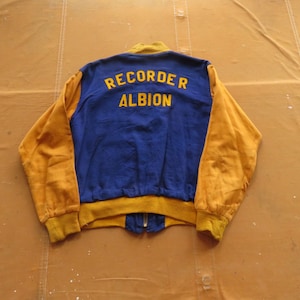 Puede incluir: Una chaqueta de béisbol azul y dorada con el texto "RECORDER ALBION" bordado en la espalda.